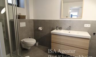 A modern bathroom with gray tiles and a bright wood vanity. It includes a shower, a toilet, and a large mirror light.