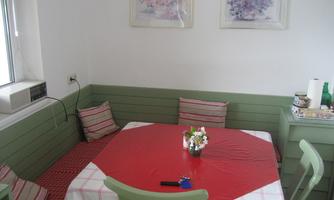 A cozy dining area with a red table and green chairs. Pictures are hanging on the wall and there are cushions on the bench.