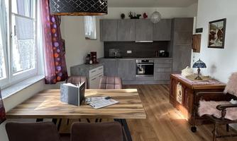 A modern living room with an open kitchen. The room is bright and welcoming, featuring a wooden dining table and cozy seating areas.