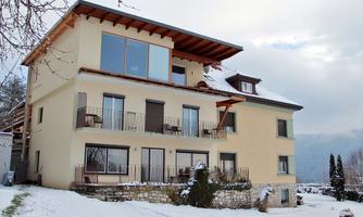 A modern house in the snow with large windows and a wooden roof. The surroundings are wintry and peaceful.