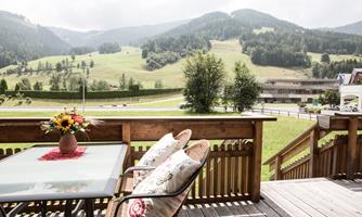 A cozy balcony with a table and chairs, surrounded by a beautiful mountain landscape. The green hills and the serene sky create a relaxed atmosphere.