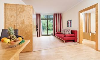 A bright living room with a red sofa and large windows overlooking a garden. Fresh fruit and a modern wooden surface are on a table.