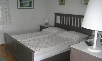 A simple bedroom with a double bed, equipped with white bedding. Some pictures hang on the walls, and there is a lamp on the nightstand.