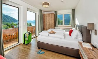 A modern room with a double bed and large windows offering a view of the mountains. Next to the bed is a green suitcase, and a couple of slippers are lying on the floor.