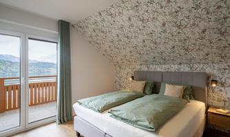 A cozy bedroom with a double bed and floral wall design. Large windows offer a beautiful view of the mountains.