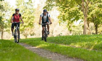 Two cyclists ride on a forest path. A green landscape with trees surrounds them.
