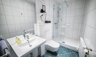 A modern bathroom with white tiles and a glass shower area. There is a sink, a toilet, and toiletries on the sink.