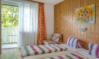 A bright room with two beds and colorful bedspreads. Large windows provide plenty of daylight and a view of a balcony.
