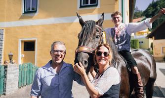 A group of three people poses with a black horse in front of a yellow house. Two people stand by the horse, while one person sits on it and smiles.