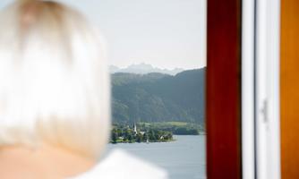 A view of a picturesque lake with a small church and surrounding mountains. In the foreground, the bright blonde hairstyle of a person is visible.
