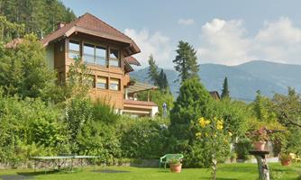 A beautiful house in the middle of a green garden. In the background, mountains and a blue sky can be seen.