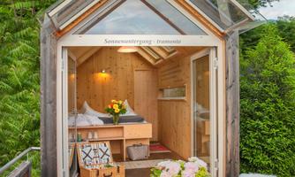 A cozy little wooden house with large windows.  
Outside is a table with chairs and fresh flowers on the table.