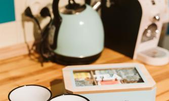 This lovely kitchen scene shows two cups on a wooden table next to a kettle and a tea box. The cups invite you to relax and enjoy.