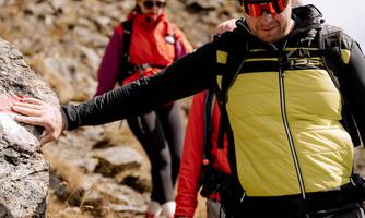A group of hikers is climbing a rocky path in the mountains. The man in front is wearing a yellow vest and sunglasses, while the others follow behind him.