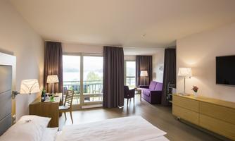 A modern hotel room with a double bed, a cozy seating area, and a view of the water. Large windows let in plenty of daylight.