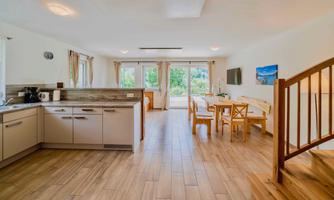 A modern, bright kitchen with a dining area. Large windows provide a view of the green surroundings.