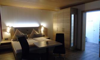 A cozy room with a large bed and a wooden ceiling. There is a table and chairs as well as access to another room.