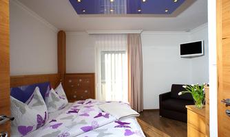 A modern bedroom with a large bed and colorful pillows. There is a television on the wall and a seating area with a sofa.