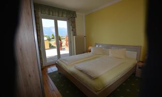 A bright bedroom with a double bed and wooden flooring. Large windows offer a view of the mountains and the balcony.