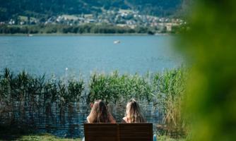 Two girls are sitting on a bench, gazing at a calm lake. In the background, green mountains and a blue sky can be seen.
