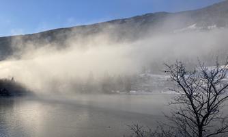 A quiet lake, surrounded by mountains, is partially shrouded in mist. The sky is clear and blue, creating a beautiful contrast to the mystical atmosphere.