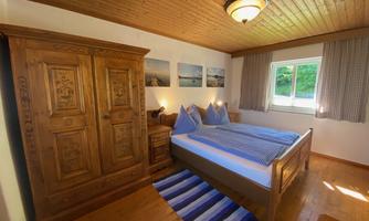 A cozy bedroom with a large wooden wardrobe and a double bed. The walls are decorated with beautiful pictures and a window provides daylight.