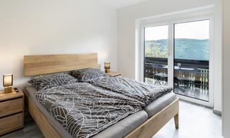 A modern bedroom with a double bed and a simple duvet. Large windows offer a view of the outside and let in plenty of daylight.