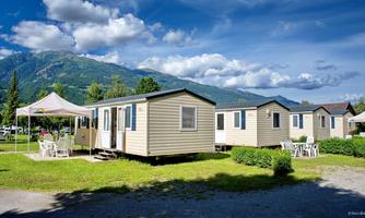 Two mobile houses in a green environment, surrounded by mountains. The sky is blue with some clouds, and there is an area with tables and chairs.