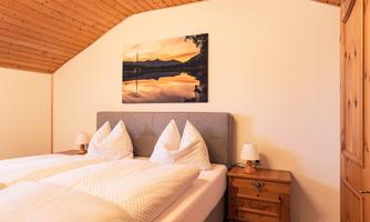 A cozy bedroom with two beds and a wooden ceiling. On the wall hangs a picture of a sunset over a lake.