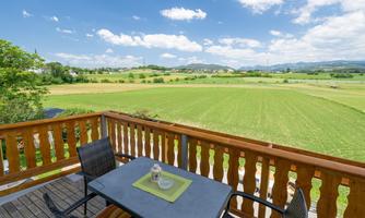 A view from a balcony over a green landscape with meadows and gentle hills. The sky is blue with some clouds.