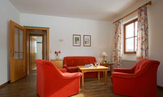 A cozy living room with red armchairs and a wooden table. The walls are decorated with pictures, and plenty of daylight comes in through the window.