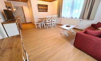 A cozy living room with wooden floors and large windows. There is a red sofa, a dining table with chairs, and a TV unit.
