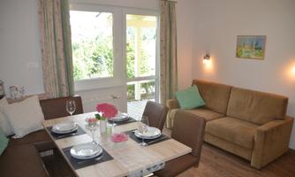 A bright living kitchen with a dining table and a cozy couch. Large windows offer a view of the greenery.
