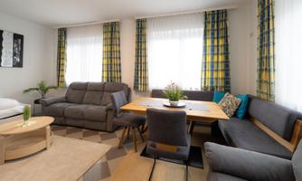 A bright living room with comfortable sofas and a dining table. Large windows let in plenty of light and the corner is decorated with plants.