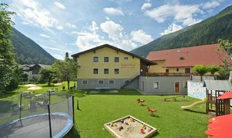 A large green property with a building in the background. There is a playground, a trampoline, and shady trees.