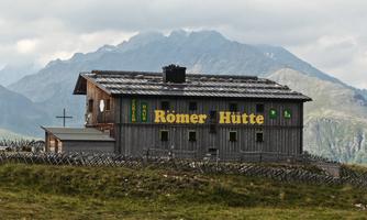 A rustic mountain cabin with the inscription "Römer Hütte" stands in an idyllic mountain landscape. In the background, majestic mountains and a cloudy sky can be seen.