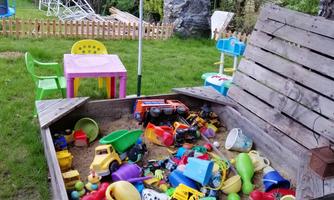 A sandbox full of colorful toys is in a garden. In the background, a table, chairs, and a swing can be seen.