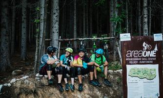 A group of mountain bikers is sitting on a log in the forest. In the background, bicycles and an information sign of the Bike Center can be seen.
