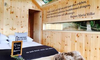 A cozy wooden house with a bed and a large window. A quote by Rainer Maria Rilke is on the wall, and a small table is decorated.