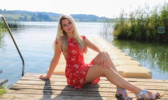 A woman is sitting smiling on a wooden jetty by the water. In the background, grassy banks and gentle hills can be seen.