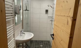 A modern bathroom with a shower, sink, and wooden ceiling. The walls are tiled white and the floor is dark gray.