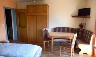 A cozy room with a double bed, a dining table, and a bench. The walls are painted in soft colors, and there is a small television on the wall.