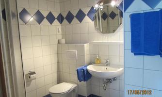 A modern bathroom with white tiles and blue accents. It features a sink, a toilet, and a mirror.