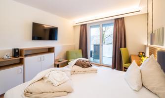 A modern hotel room with a round bed, a television, and a small table. Large windows let in plenty of daylight and provide access to a balcony.
