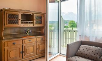 A cozy living area with a wooden cabinet and a couch. The balcony doors are open and offer a view of the green landscape.
