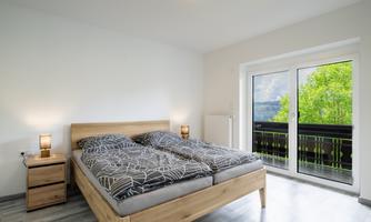 A modern bedroom with a double bed and two nightstands. Large windows offer a view of a green balcony.