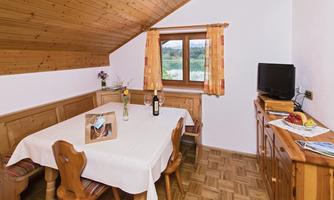 A cozy dining area with a wooden ceiling and a table surrounded by chairs. A window with curtains lets daylight in, and a small television is on the sideboard.