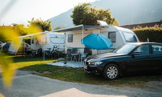 A motorhome park with two vehicles and a blue sun umbrella. In the background, there are mountains and trees.