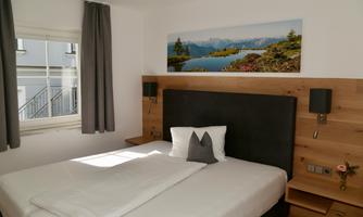 A bright bedroom with a large bed and wooden details. On the wall hangs a picture of a landscape with mountains and a lake.