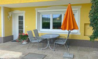 A cozy outdoor area with a round table and four chairs. An orange umbrella stands next to it in front of a yellow wall.